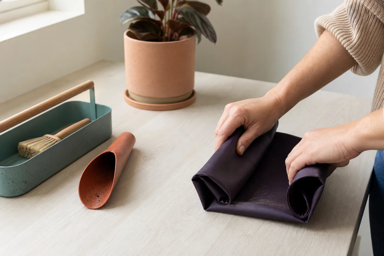 Hands folding a repotting mat beside compact plant-care tools on a small work surface.